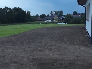 Wyrównany teren pod trawnik przy domu jednorodzinnym, świeżo rozplantowana ciemna ziemia, w tle zabudowa wiejska i zieleń drzew, widoczne ślady po pracy maszyn.