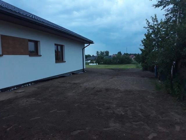 Przygotowany teren pod trawnik przy nowym budynku z widoczną czarną folią hydroizolacyjną przy fundamencie i świeżo wyrównaną ziemią w pochmurny dzień.