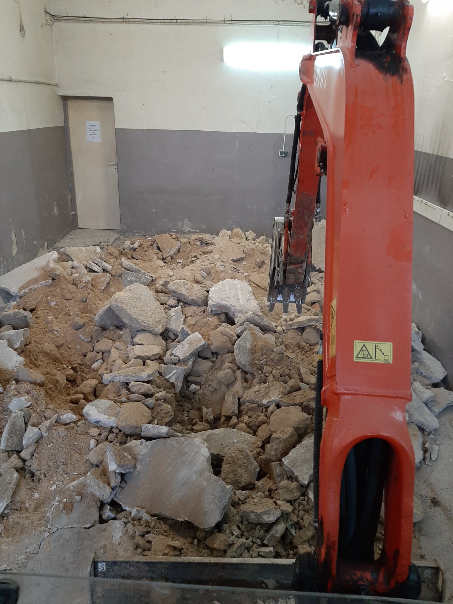 Pomarańczowa minikoparka w trakcie rozbiórki posadzki wewnątrz pomieszczenia, gruz i piasek na podłodze, widoczne ściany w dwóch odcieniach szarości, drzwi w tle.