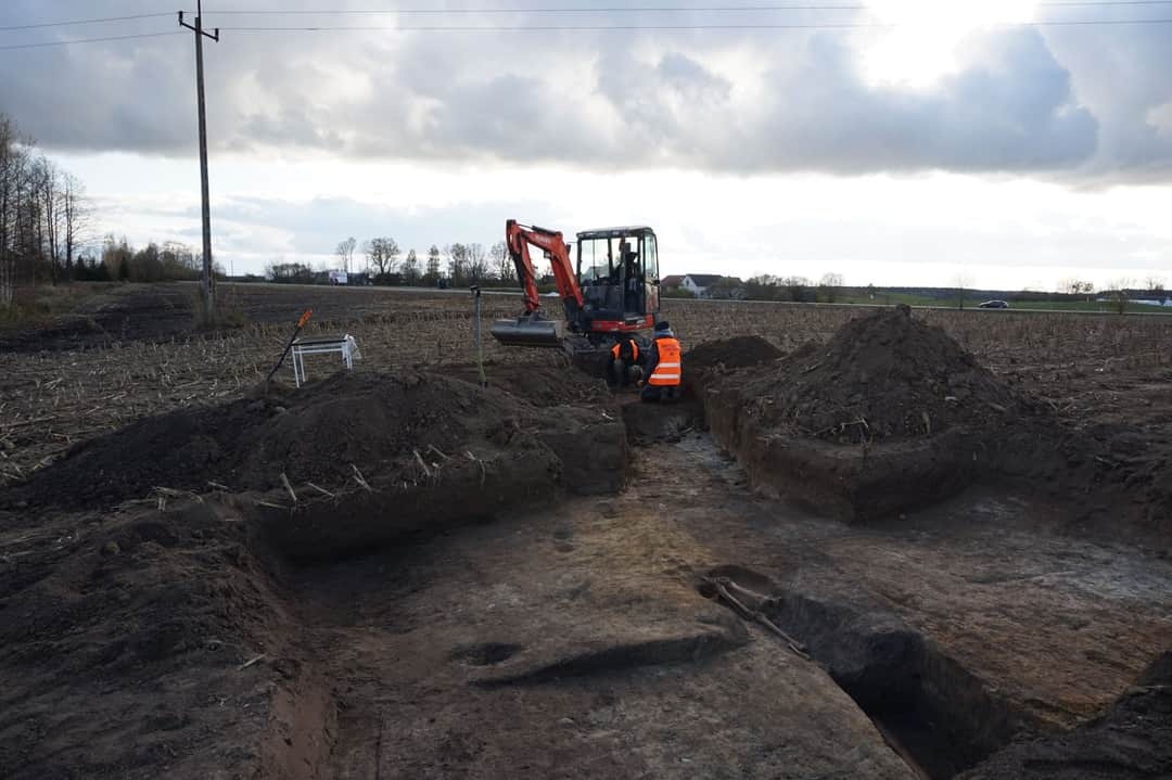 Mała koparka Kubota i dwóch pracowników w pomarańczowych kamizelkach wykonuje wykop liniowy w polu z resztkami pożniwnymi, widoczne pryzmy ziemi i pochmurne niebo.