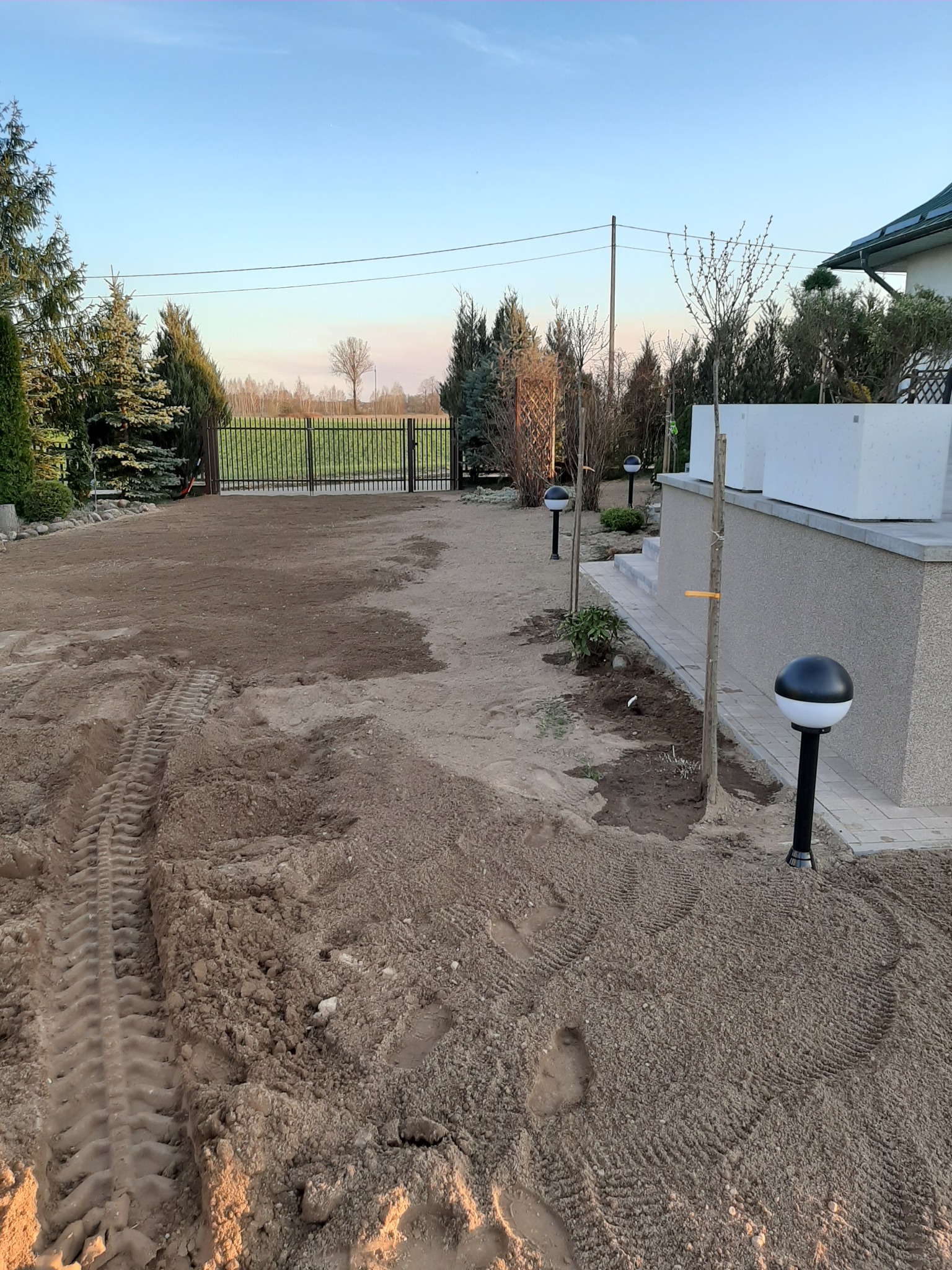 Przygotowany teren pod trawnik z widocznymi śladami opon i butów, w tle ogrodzenie posesji i drzewa iglaste, oświetlenie ogrodowe wzdłuż ścieżki.