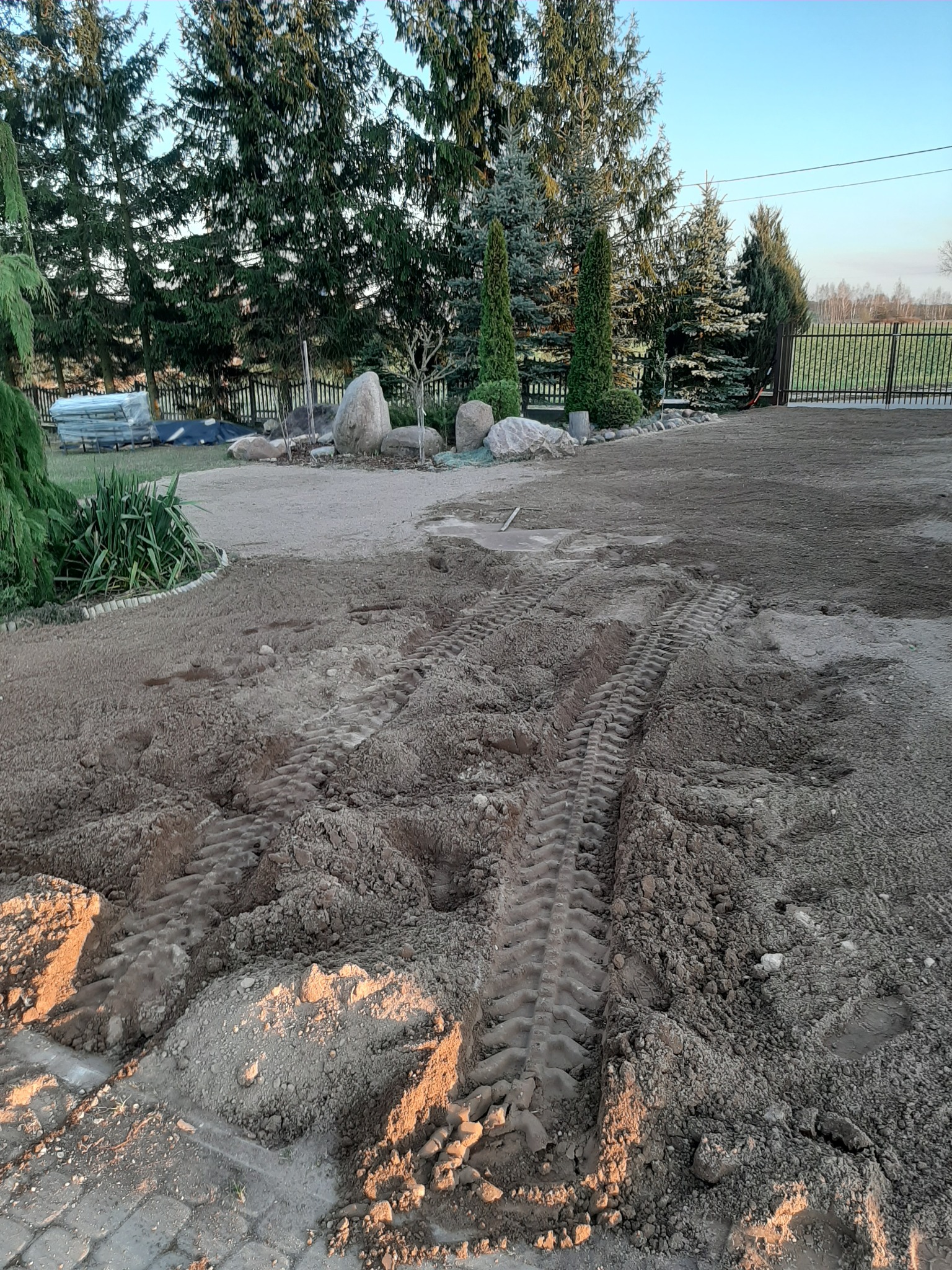 Przygotowany teren pod budowę z widocznymi śladami opon ciężkiego sprzętu i częściowo ułożoną kostką brukową na obrzeżach, w tle drzewa iglaste i ogrodzenie.