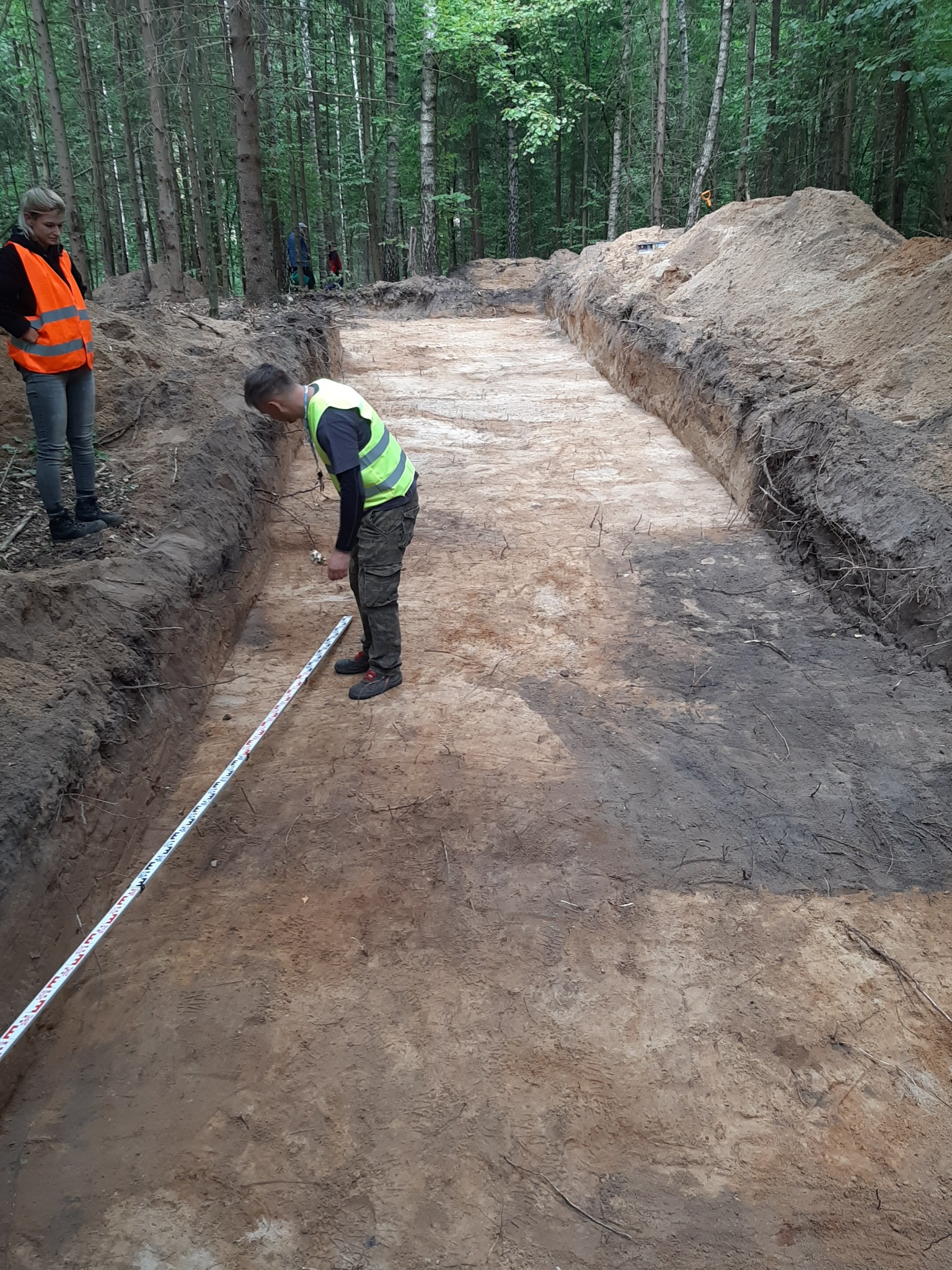 Wykonawca mierzy długość wykopu w lesie, pracownicy w odblaskowych kamizelkach nadzorują prace ziemne.