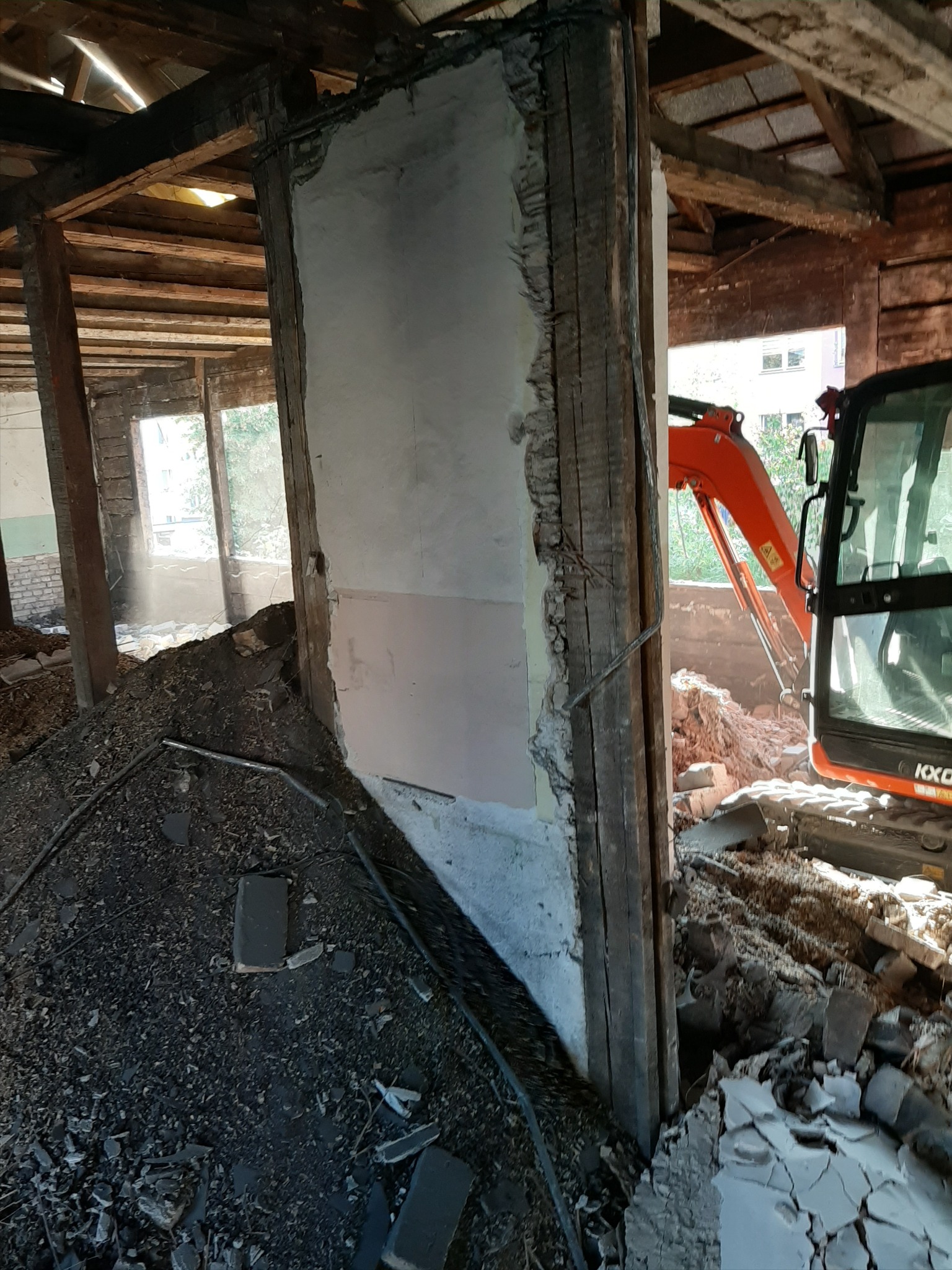 Wnętrze budynku w trakcie rozbiórki, widoczna ściana z fragmentami tynku i odsłoniętą konstrukcją drewnianą, gruzowisko na podłodze oraz pomarańczowa koparka w tle, pracująca przy usuwaniu...