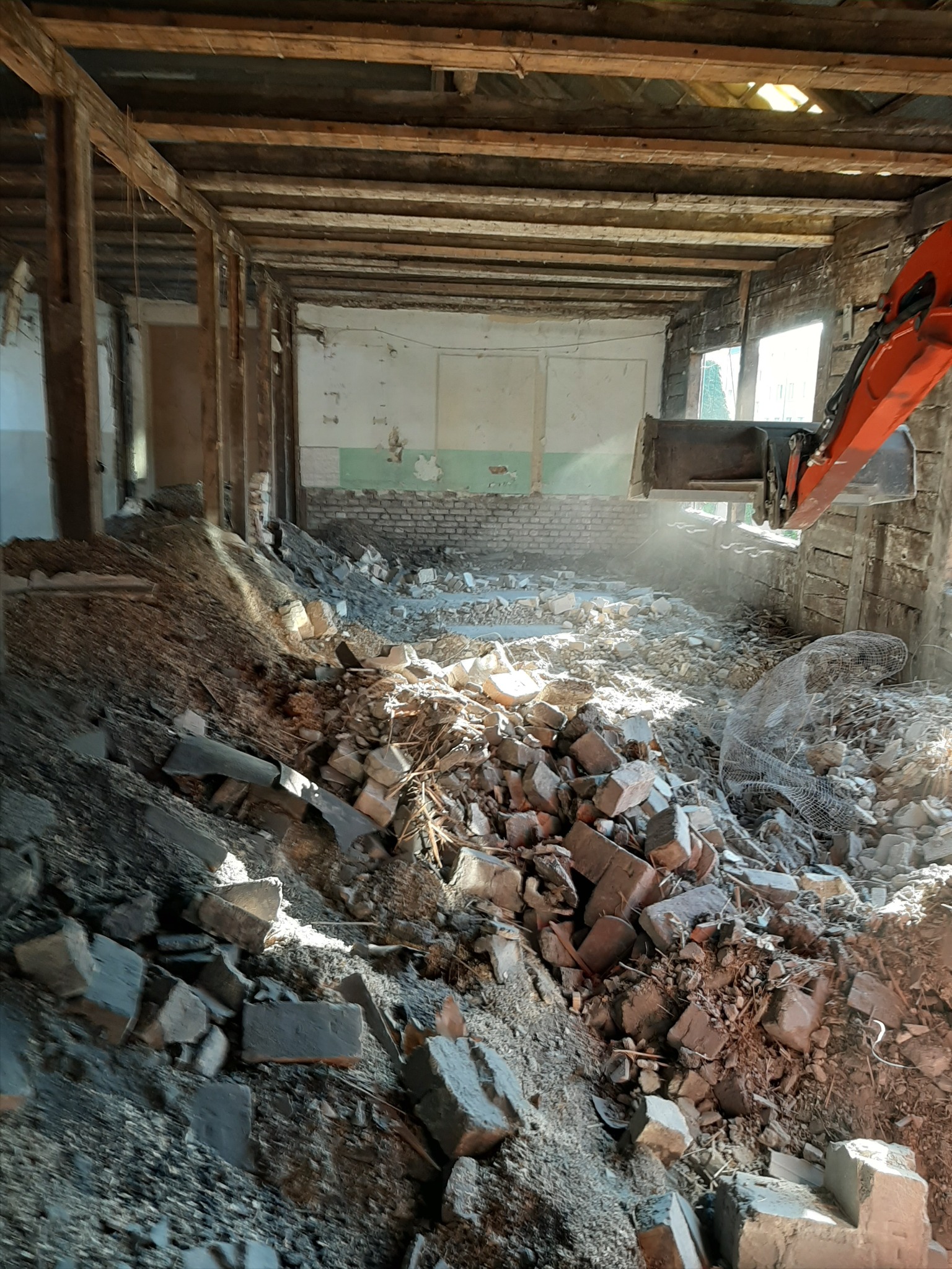 Wnętrze budynku w trakcie rozbiórki, widoczny gruz, cegły i fragment pomarańczowego ramienia koparki, drewniana konstrukcja dachu częściowo usunięta, w tle okna w zdemolowanej ścianie.