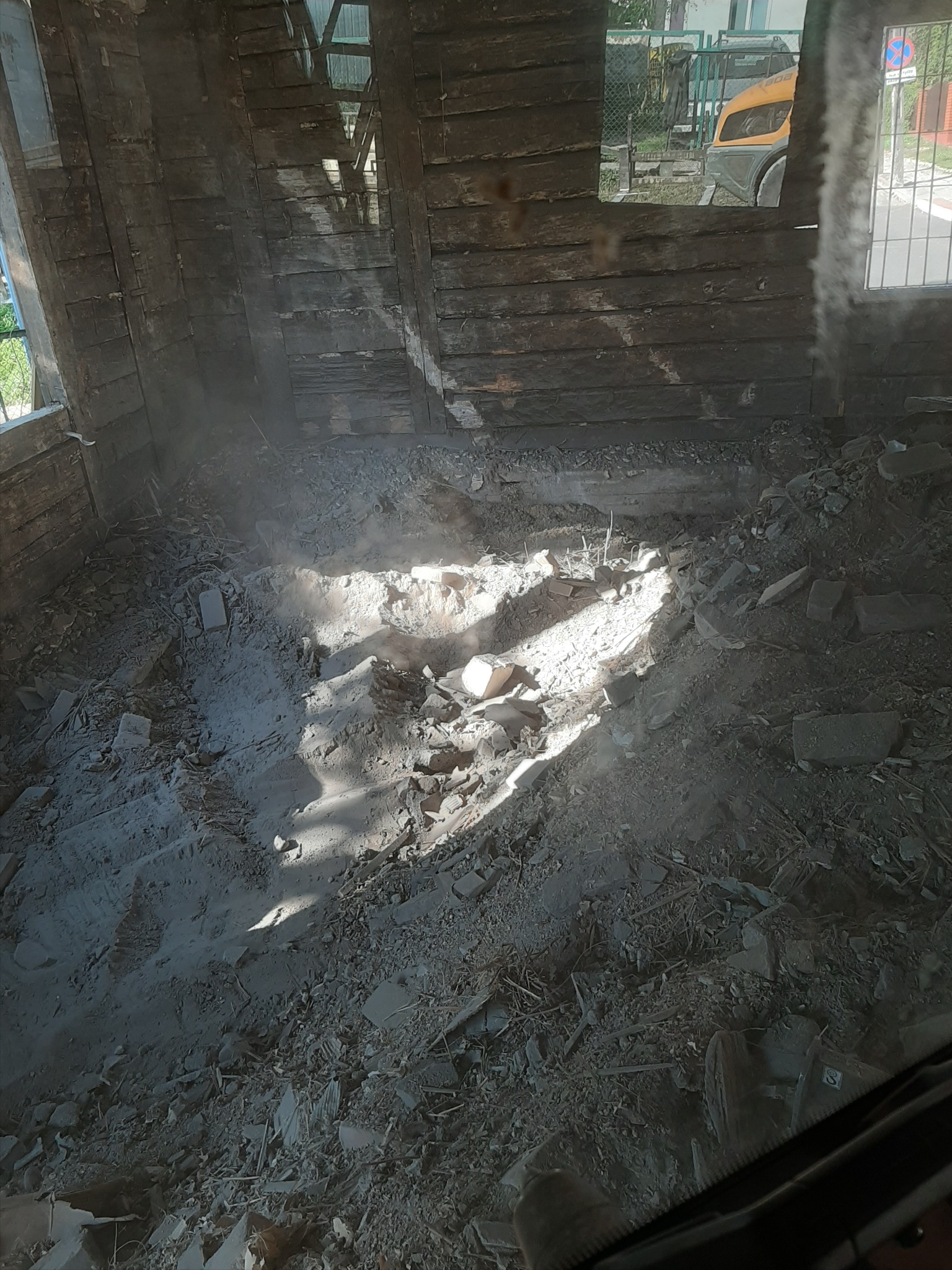 Wnętrze starego budynku w trakcie rozbiórki, widoczne drewniane ściany i gruz na podłodze, z przebijającym się światłem słonecznym tworzącym plamy na podłożu. W tle, przez okno, widoczny fragment...