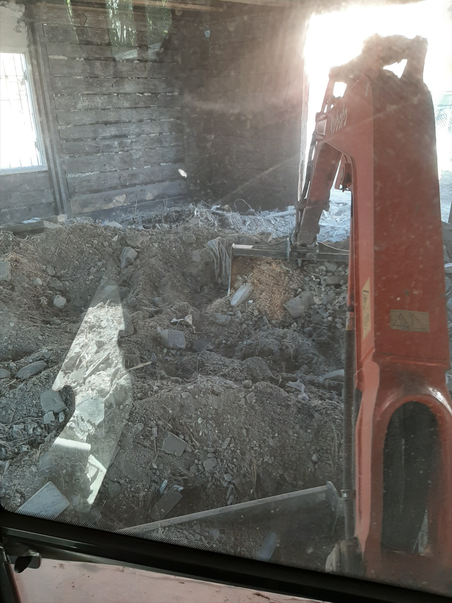Wnętrze budynku w trakcie rozbiórki widziane z kabiny koparki, z widoczną ścianą z drewnianych bali i gruzem.