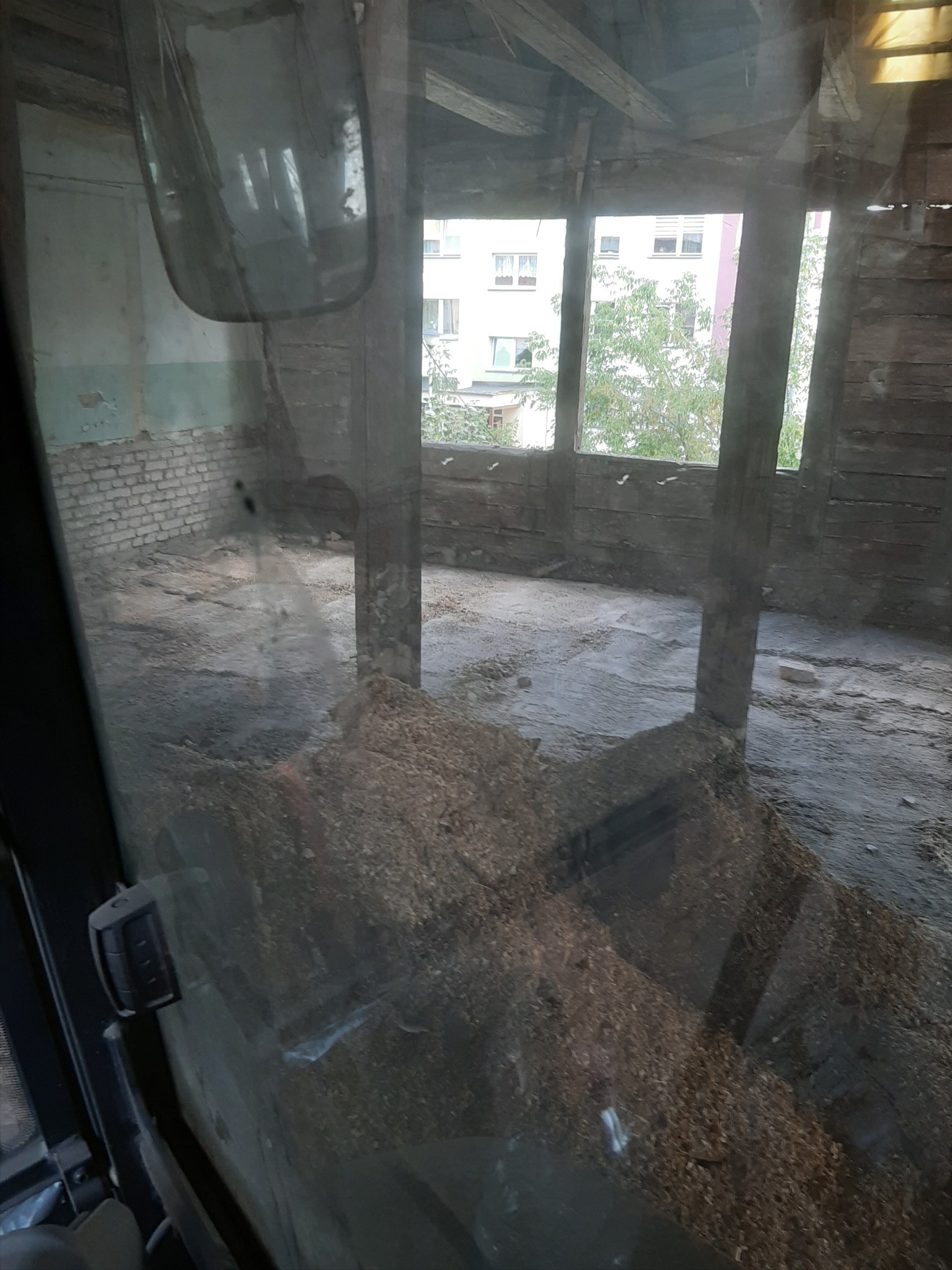 Wnętrze budynku w trakcie rozbiórki, widok z kabiny maszyny budowlanej na stertę gruzu i odsłonięte ściany z cegły i drewna, okna wychodzące na budynek mieszkalny.