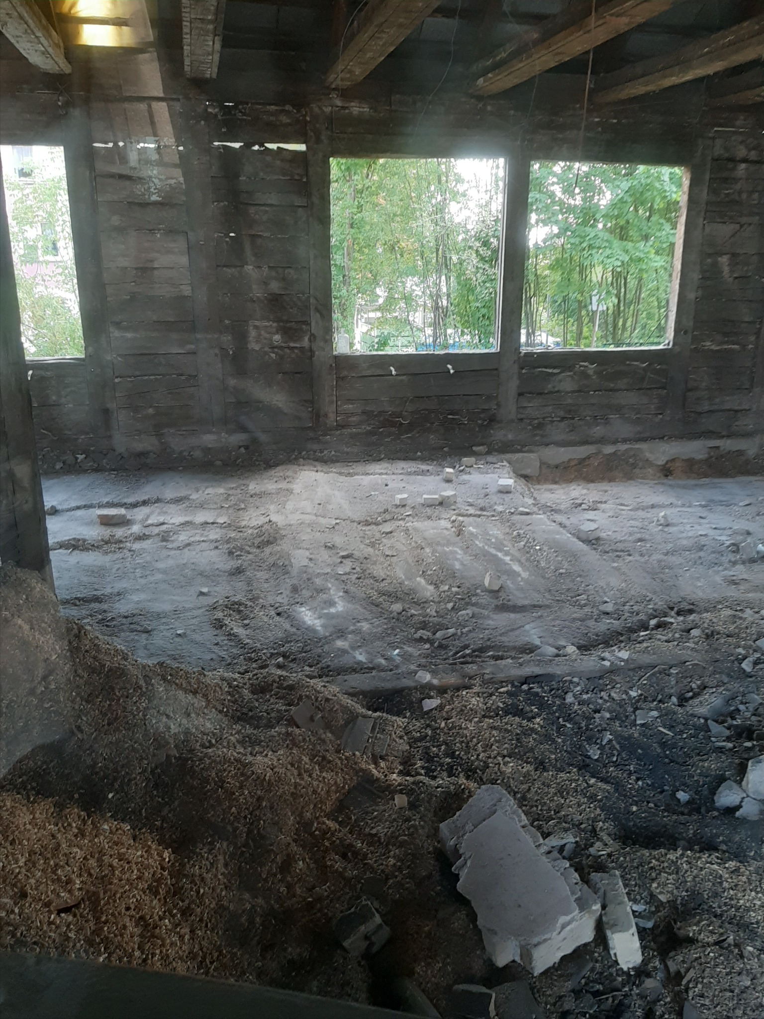 Wnętrze budynku w trakcie rozbiórki, widoczne drewniane elementy konstrukcyjne, okna bez szyb z widokiem na zieleń, gruz i resztki materiałów budowlanych na podłodze.