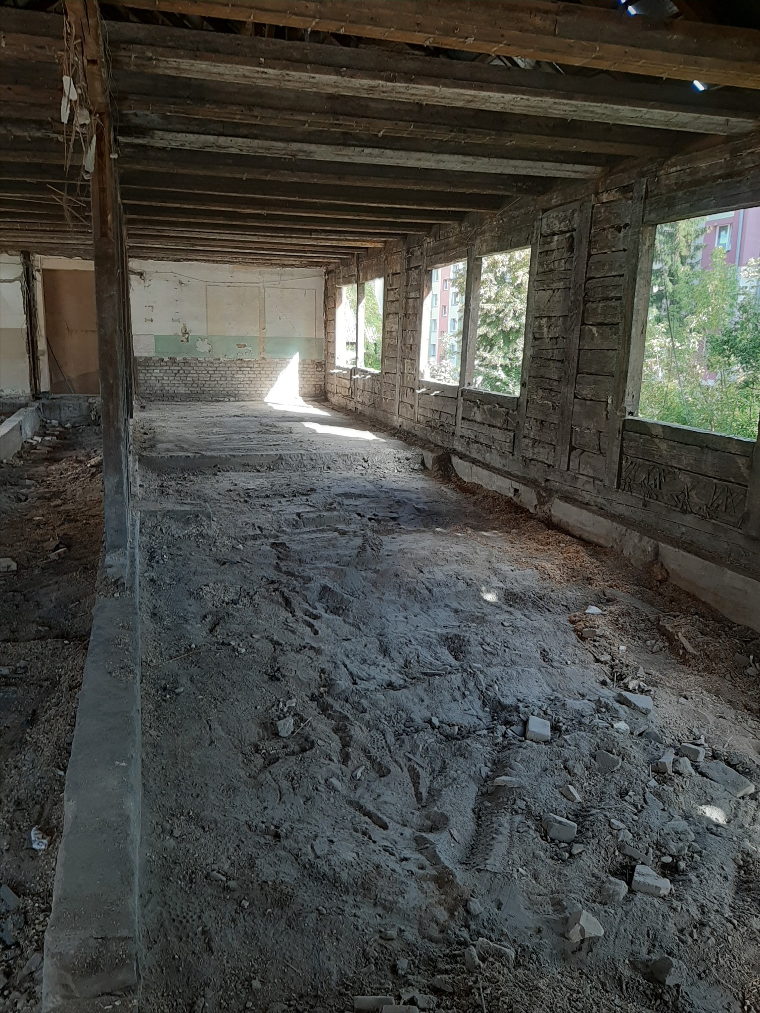 Wnętrze budynku w trakcie rozbiórki, widoczne drewniane belki stropowe, ślady po ścianach i okna z widokiem na zieleń.