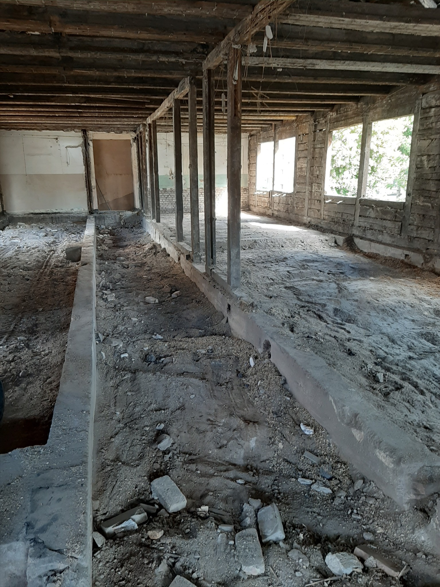 Wnętrze budynku w trakcie rozbiórki, widoczne drewniane elementy konstrukcyjne, resztki ścian i gruz na podłodze, światło dzienne wpada przez okna.