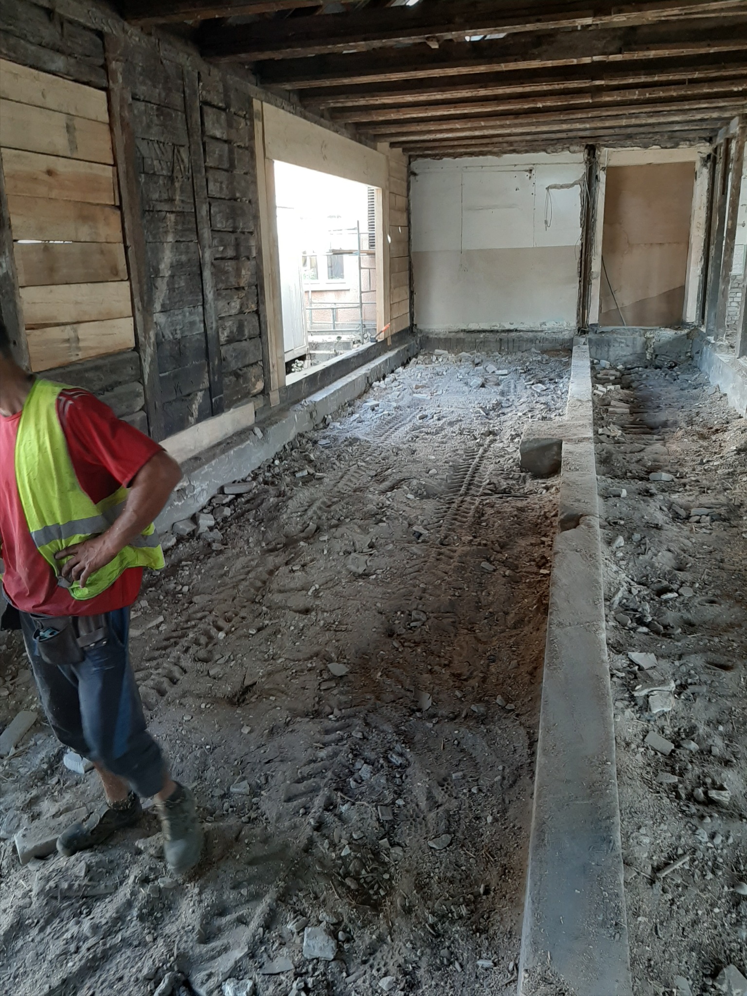 Wnętrze budynku w trakcie rozbiórki, widoczne odsłonięte fundamenty, gruz i ślady opon na ziemi, pracownik w odblaskowej kamizelce w tle.
