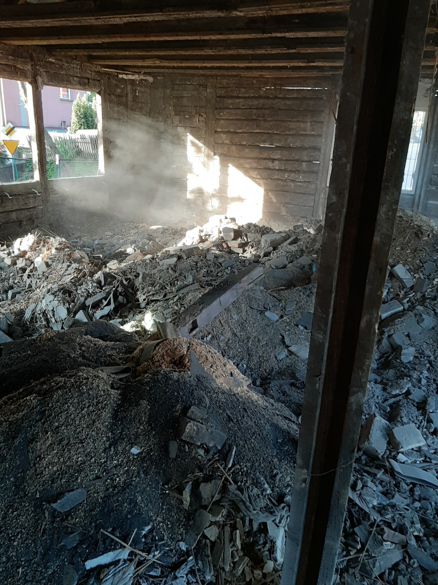 Wnętrze zrujnowanego budynku z drewnianą konstrukcją, gruzowisko i pył wpadający przez okna tworzą promienie światła.