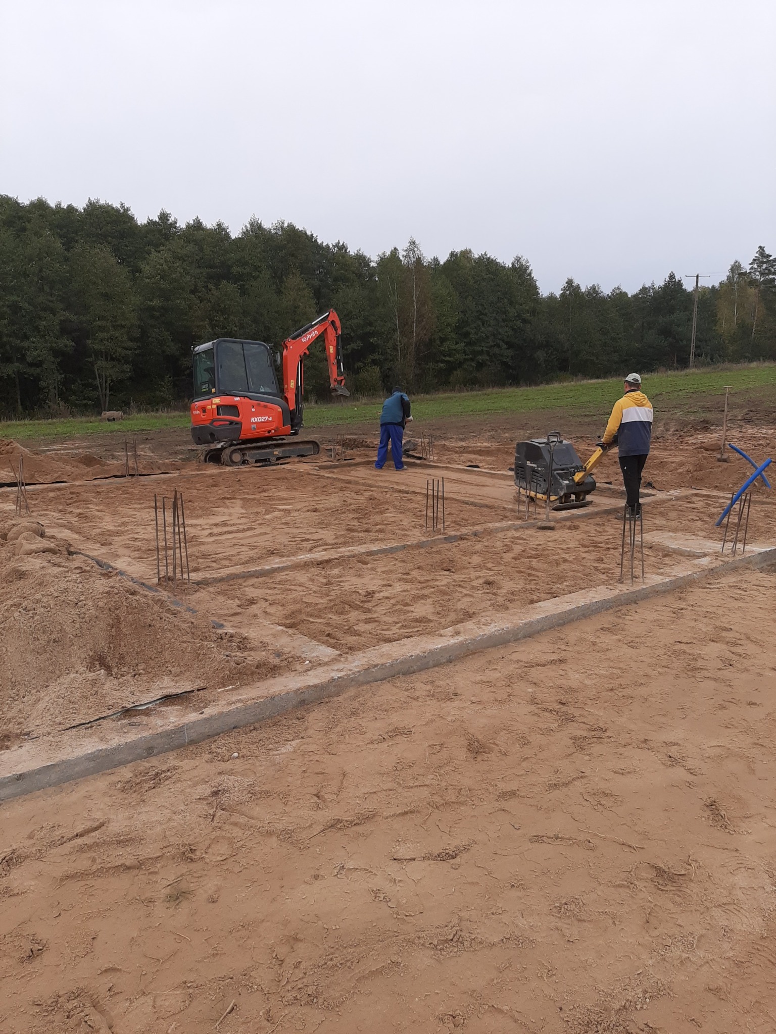 Wykop pod fundamenty z widocznym szalunkiem, wystającymi prętami zbrojeniowymi i zagęszczarką wibracyjną w trakcie pracy oraz minikoparką w tle.