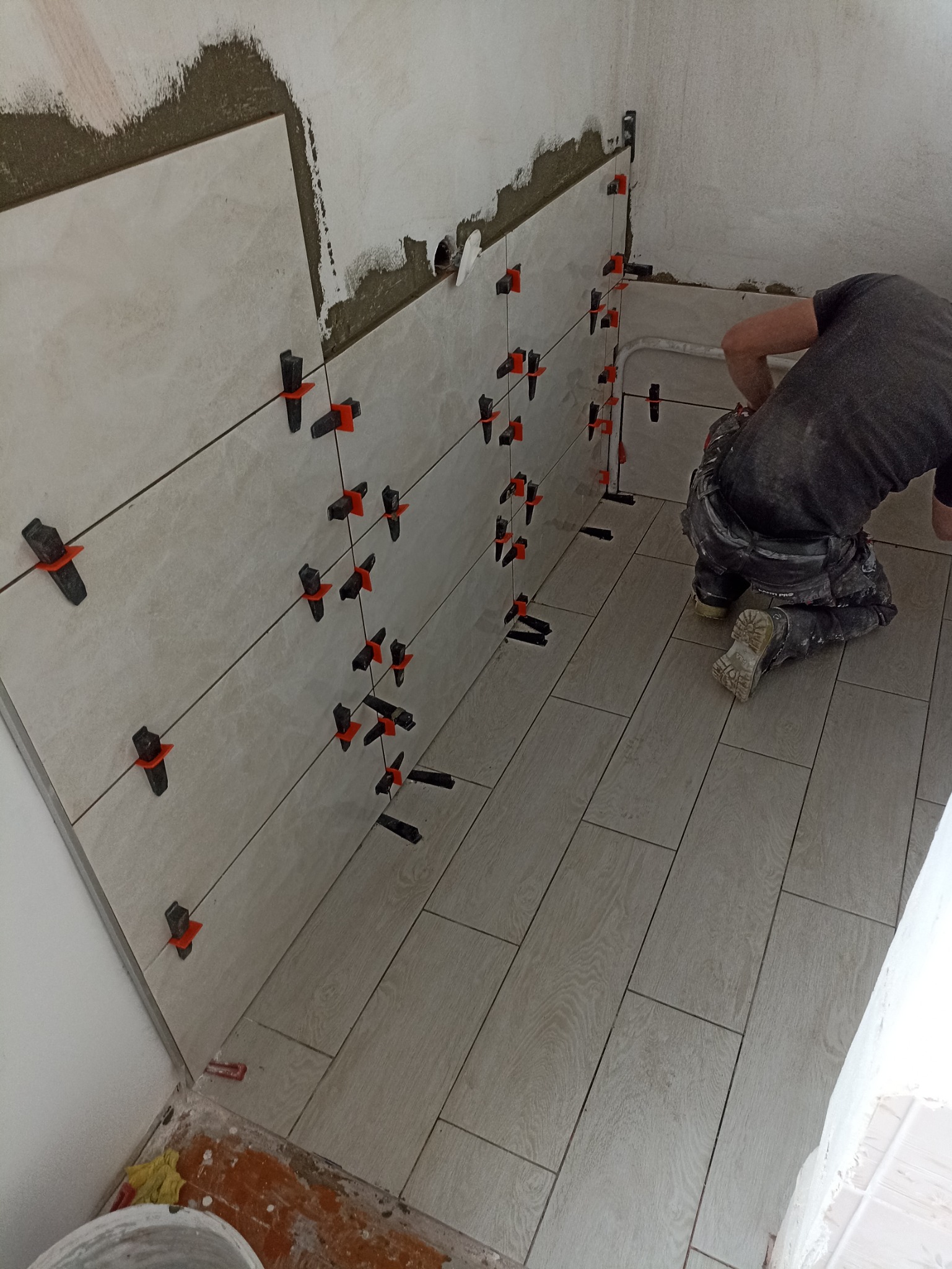 Układanie płytek podłogowych i ściennych w łazience, z użyciem klipsów poziomujących, widoczne niedokończone fragmenty ściany.