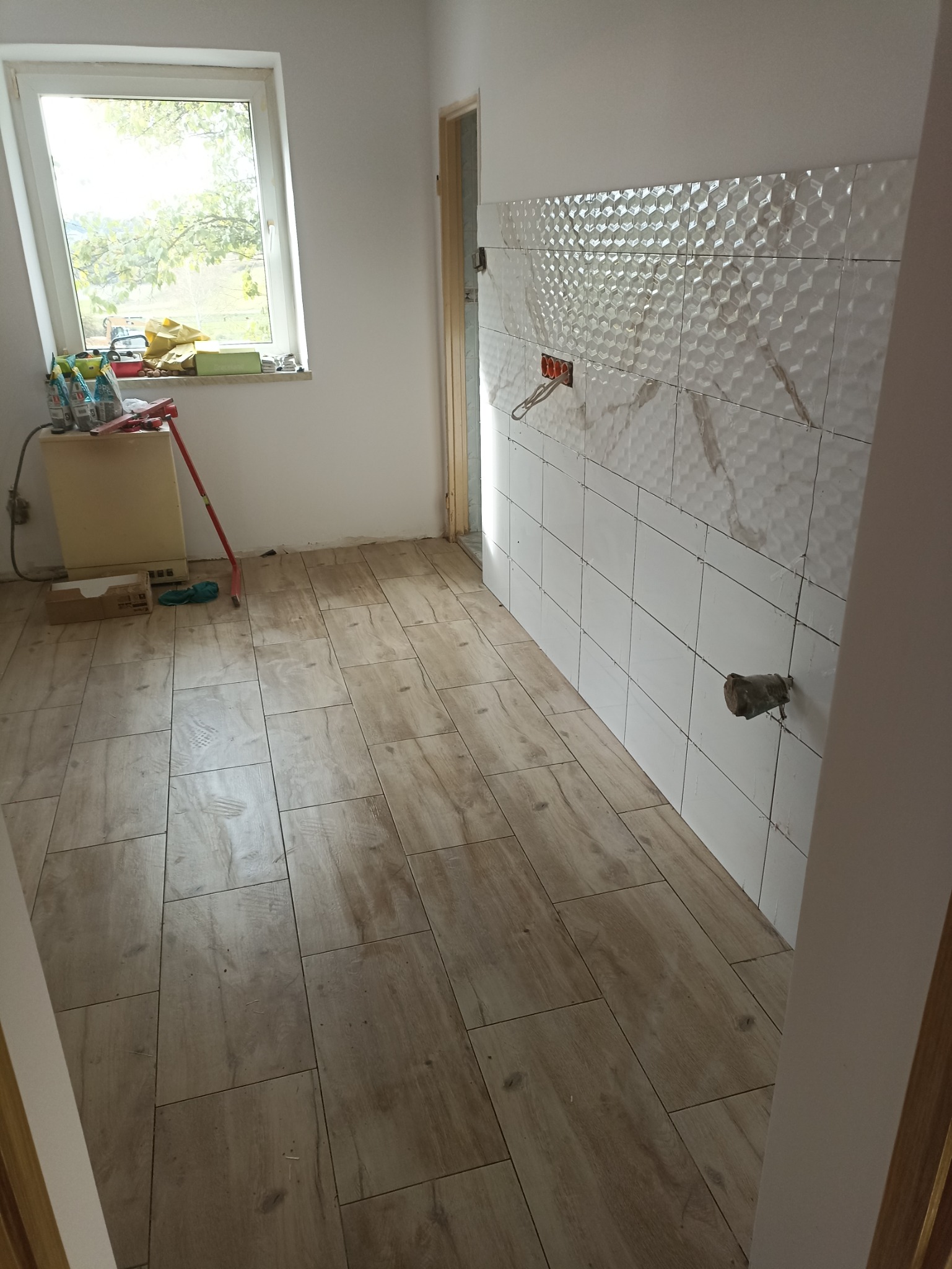 Wnętrze kuchni w trakcie prac wykończeniowych, widoczne ułożone płytki podłogowe imitujące drewno, częściowo wykończona ściana z białych płytek o trójwymiarowej strukturze, wystające kable...