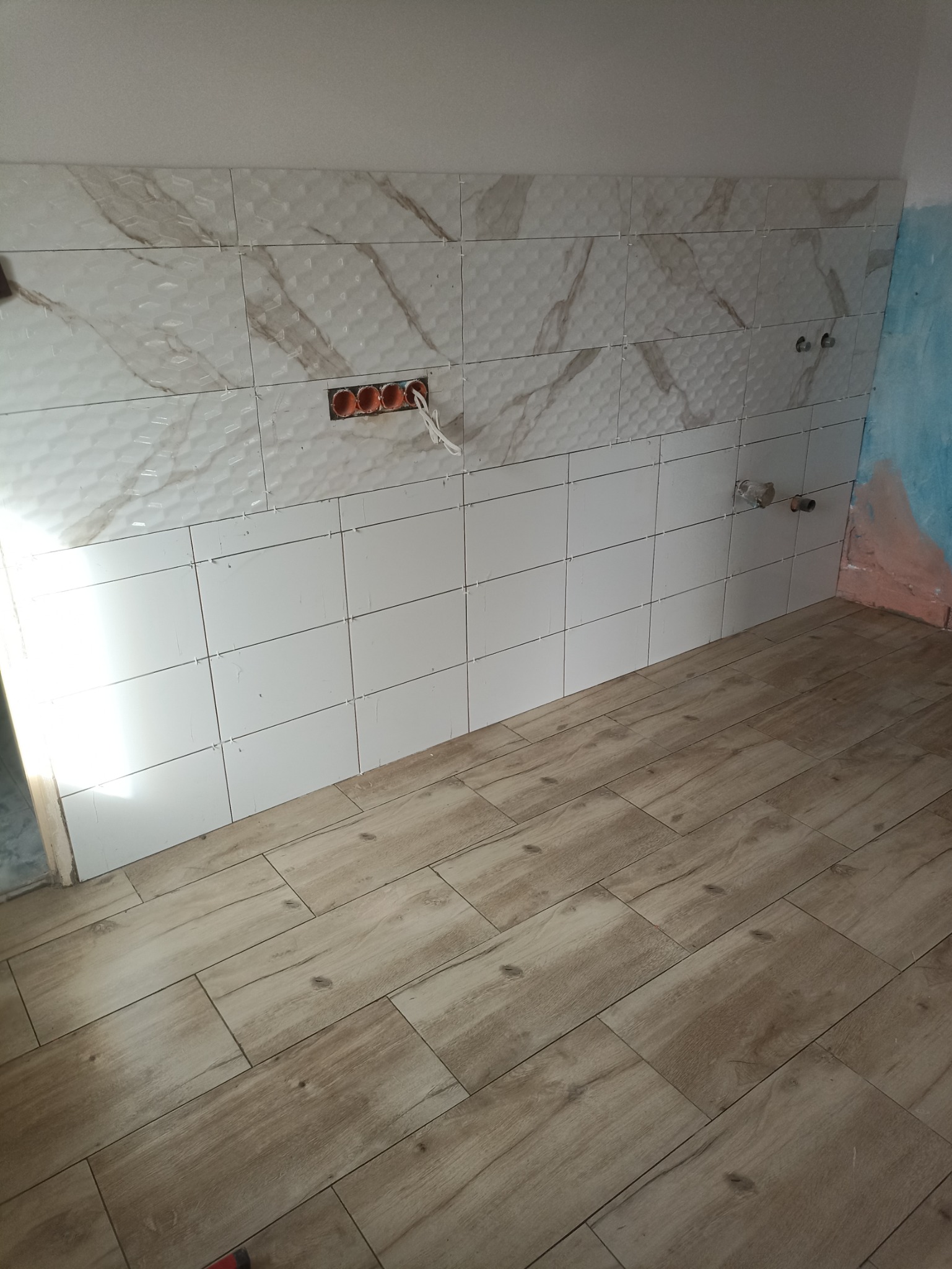 Fragment kuchni w trakcie wykańczania: częściowo ułożone białe płytki ścienne z beżowym wzorem, widoczne gniazdka elektryczne i rury doprowadzające wodę, na podłodze płytki imitujące drewno.