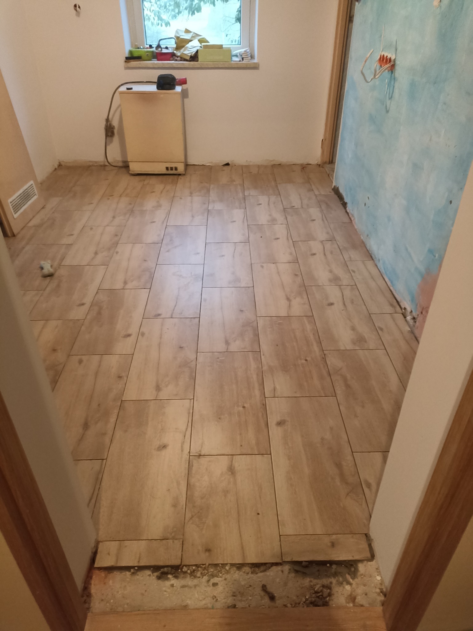 Ułożona podłoga z płytek imitujących drewno w trakcie remontu łazienki, widoczne niedokończone ściany i instalacja elektryczna.