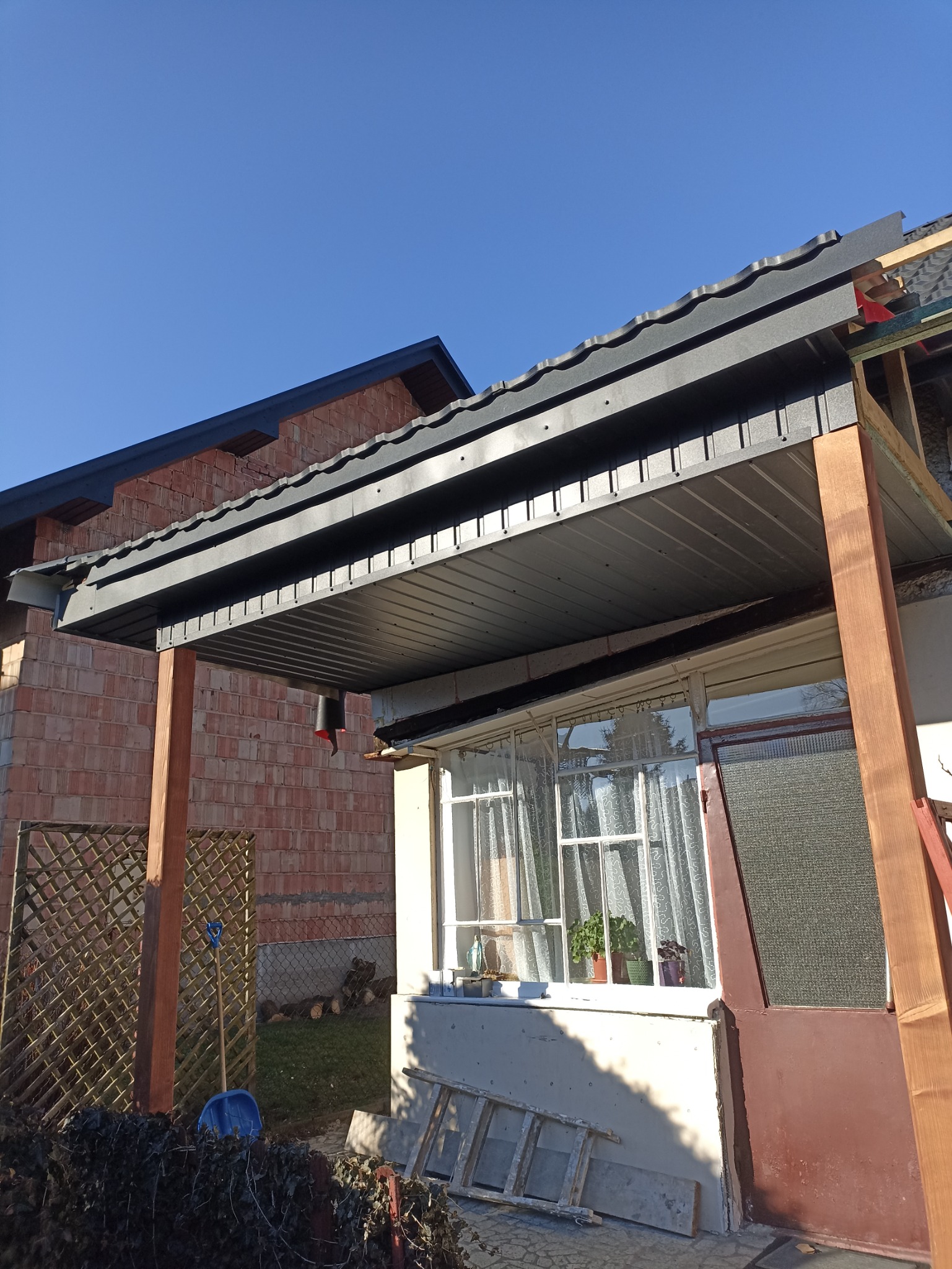 Montaż zadaszenia tarasu z blachy trapezowej w kolorze grafitowym, widoczne drewniane słupy konstrukcyjne, okno z firankami i doniczkami, ceglany budynek w tle, niebieskie niebo.