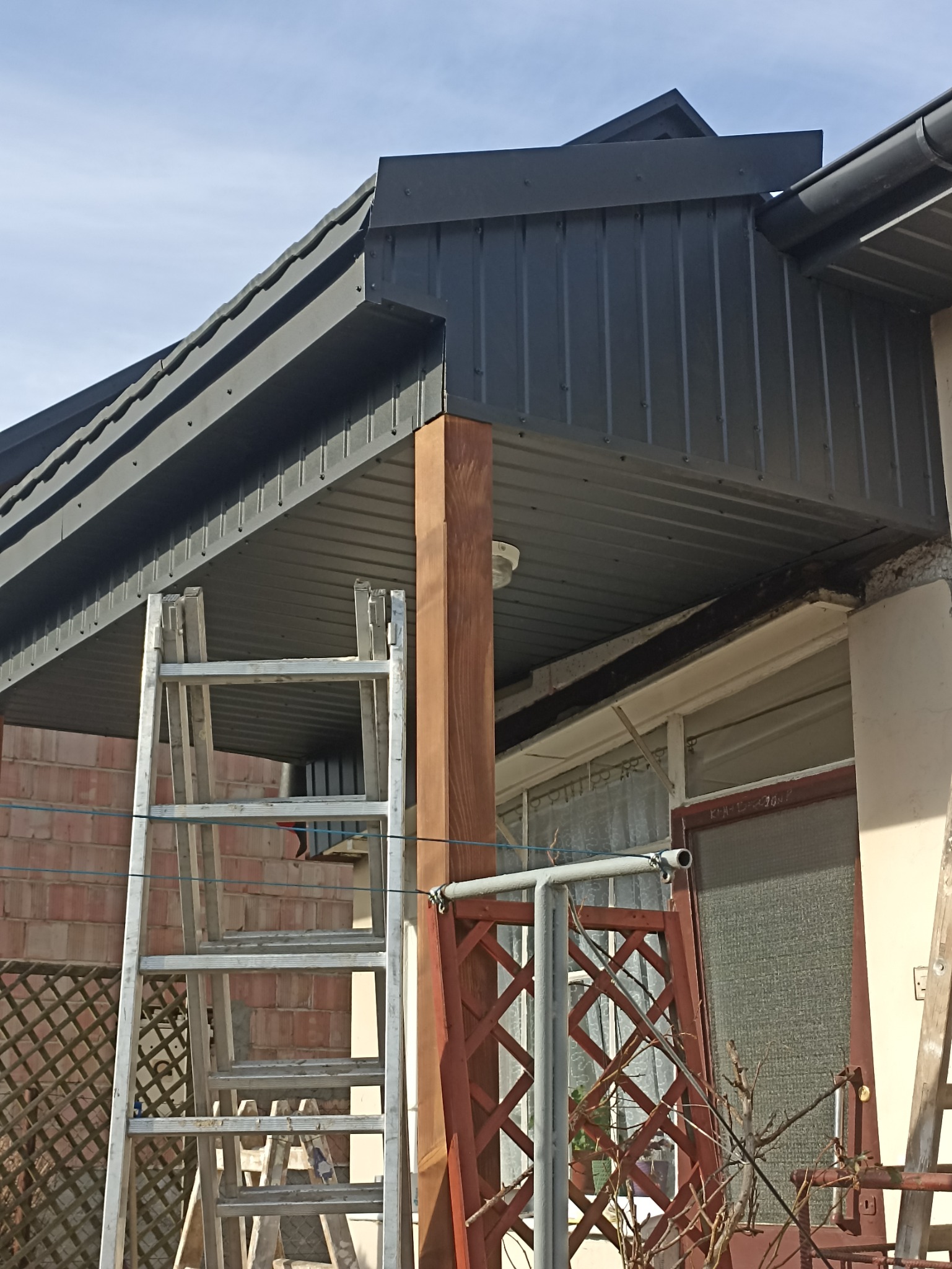 Montaż podbitki dachowej z blachy trapezowej, widoczna drabina oparta o ścianę budynku, drewniana kolumna podtrzymująca konstrukcję dachu, oraz fragment ściany z oknem.
