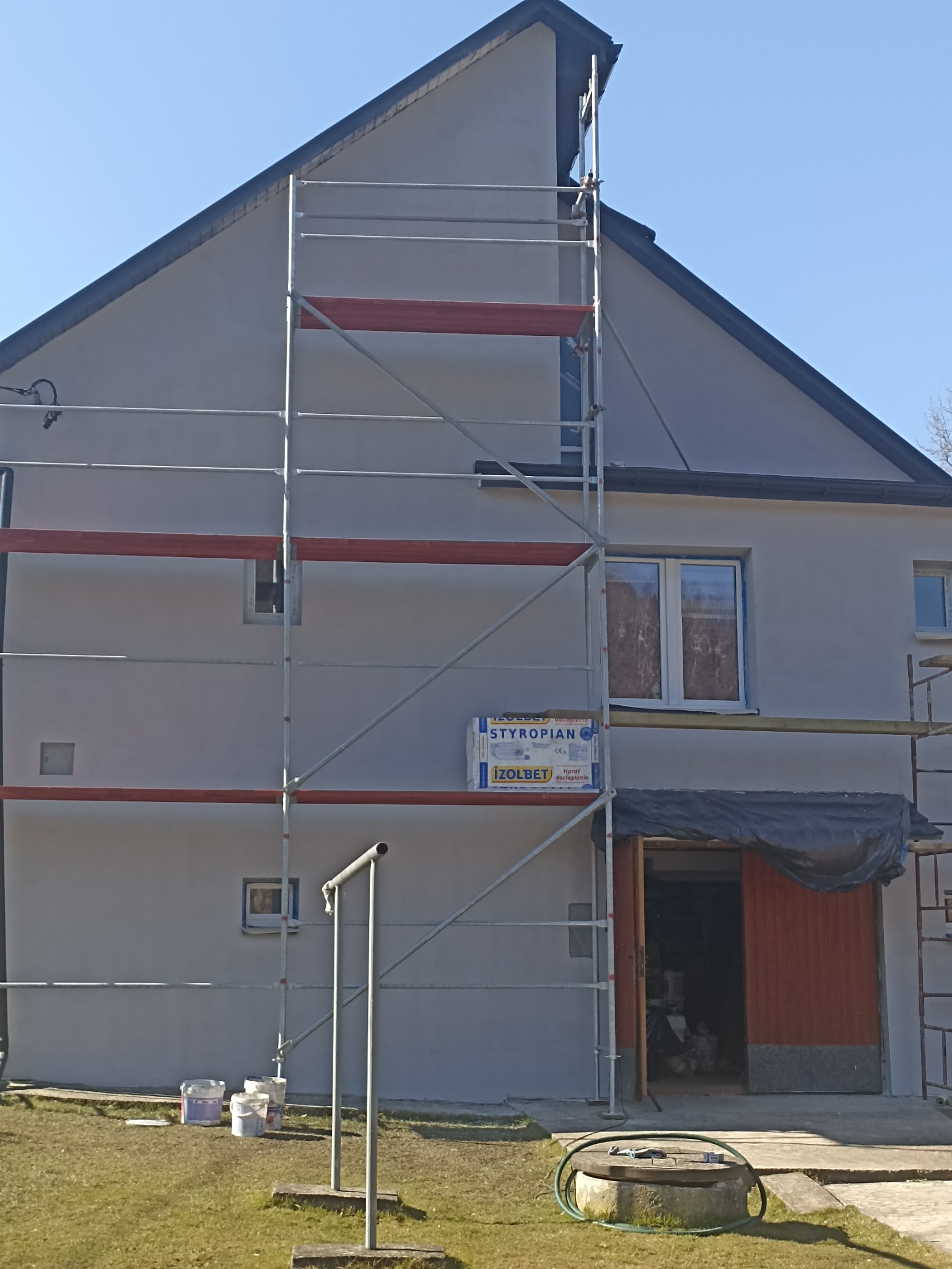 Ocieplanie ścian zewnętrznych domu jednorodzinnego z użyciem styropianu Izolbet, widoczne rusztowanie i puszki po farbie na trawie.
