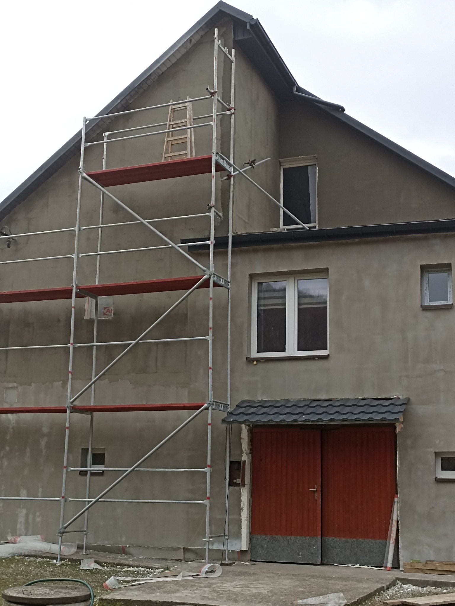 Elewacja domu w trakcie ocieplania, widoczne rusztowanie z drewnianą drabiną, ściany pokryte szarym tynkiem, częściowo nieocieplone, okna w białych ramach.