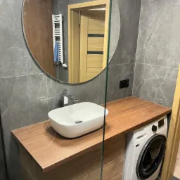 Nowoczesna łazienka z szarymi płytkami, drewnianym blatem pod umywalką, okrągłym lustrem i pralką Samsung.