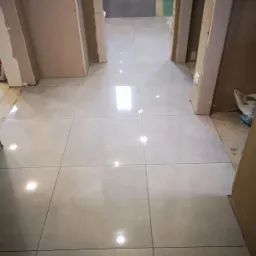 Wyłożona kremowymi płytkami podłoga w trakcie remontu, widoczne surowe ościeżnice drzwiowe i fragmenty szarej ściany w głębi.