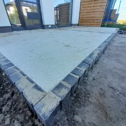 Nowo ułożony taras z jasnych płyt ograniczonych ciemnoszarą kostką brukową, widoczny kabel zasilający i gniazdko elektryczne w pobliżu, ziemia wokół tarasu przygotowana pod dalsze prace.