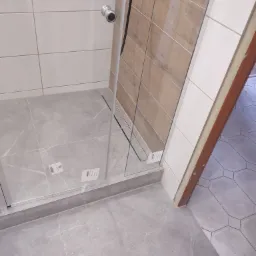 Wykończona kabina prysznicowa z szarymi płytkami imitującymi marmur na podłodze i częściowo na ścianach, z widocznym odpływem liniowym i fragmentem drzwi do łazienki z widoczną inną, wzorzystą...