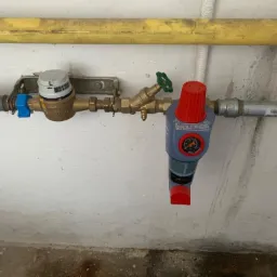 Szare rury wodne z zamontowanym licznikiem, zaworem z zielonym pokrętłem i filtrem z czerwonym regulatorem ciśnienia na tle białej ściany.