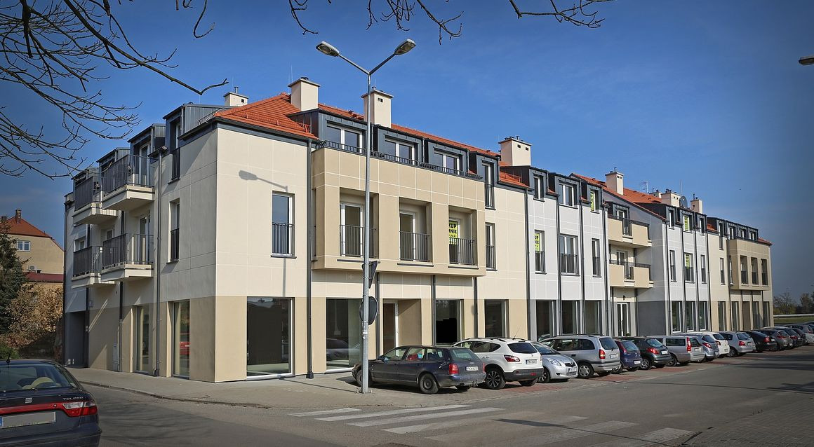 Nowoczesny budynek wielorodzinny z balkonami i podziemnym parkingiem, wykończony w jasnych kolorach, widoczny z perspektywy ulicy z zaparkowanymi samochodami.