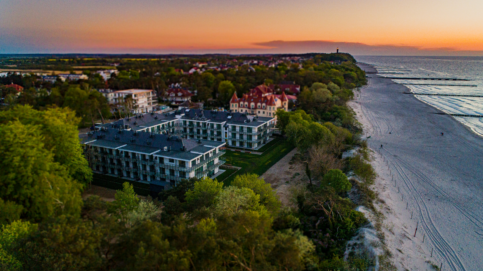 Widok z lotu ptaka na kompleks apartamentów w nadmorskiej miejscowości o zmierzchu, z widoczną plażą, lasem i latarnią morską w oddali.