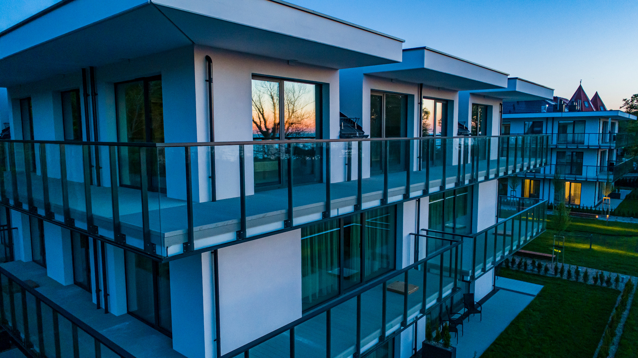 Nowoczesny budynek wielorodzinny z balkonami o szklanych balustradach, widok z góry o zmierzchu, odbicie drzewa w oknach.