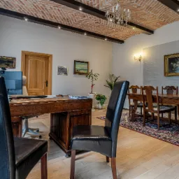 Wnętrze kancelarii z biurkiem, krzesłami, drewnianym stołem konferencyjnym i ceglanym sufitem ze sztukaterią, oświetlone lampami i żyrandolem.