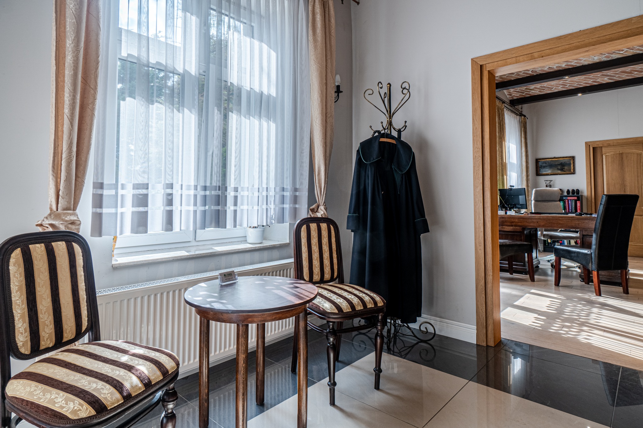 Eleganckie wnętrze kancelarii w Gdańsku: dwa krzesła ze stolikiem przy oknie z zasłonami, w tle wieszak z togą i widok na biuro z biurkiem i krzesłami.