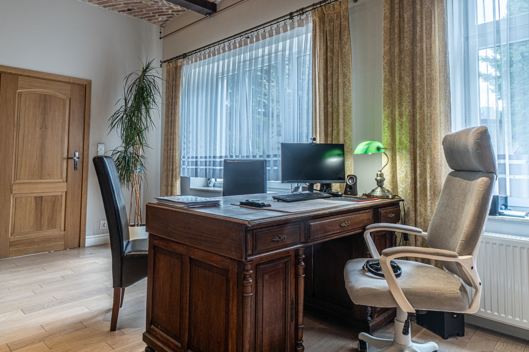 Gabinet z drewnianym biurkiem, komputerem, lampką, dwoma krzesłami i oknem z zasłonami w tle.