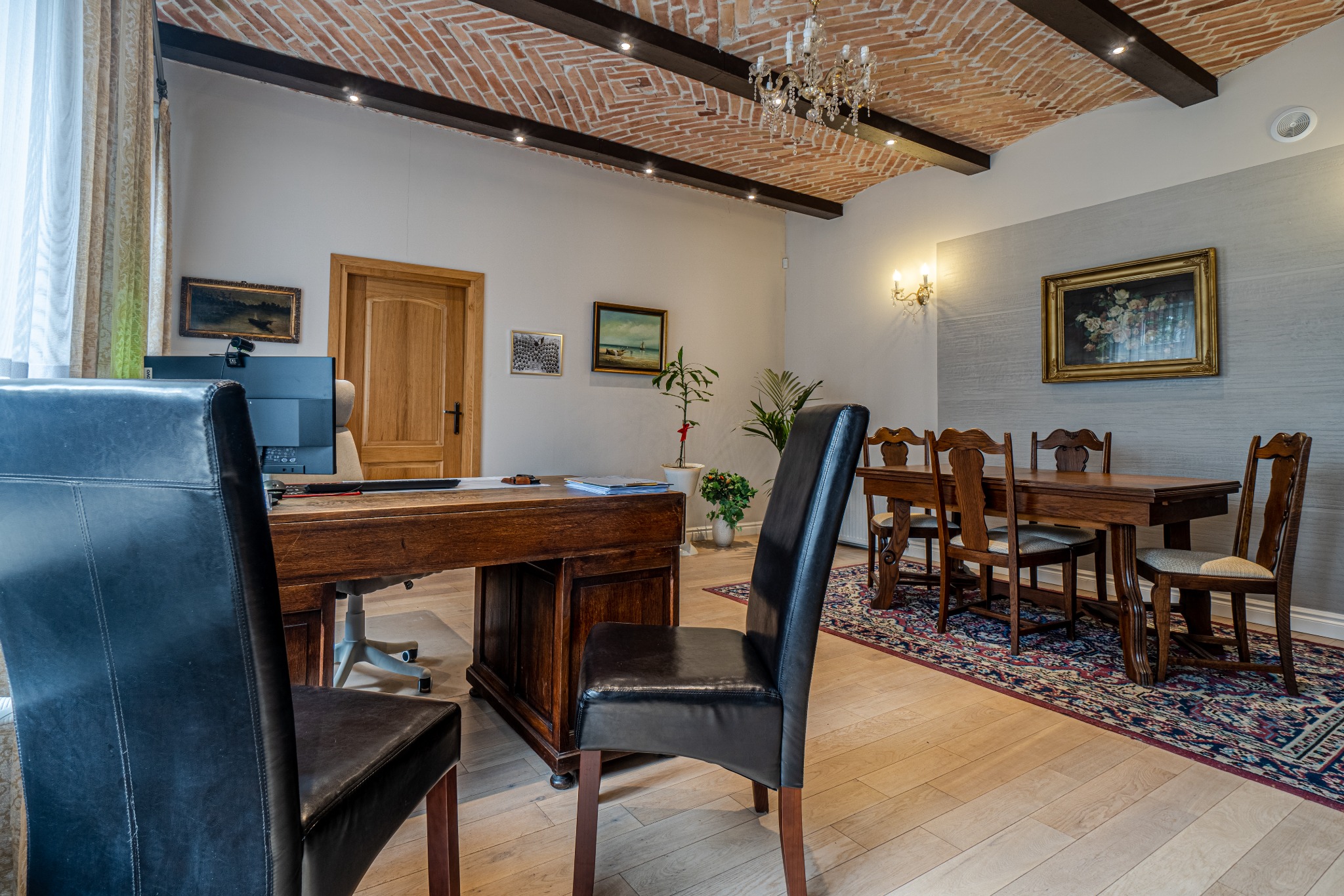 Wnętrze kancelarii z biurkiem, krzesłami, drewnianym stołem konferencyjnym i ceglanym sufitem ze sztukaterią, oświetlone lampami i żyrandolem.