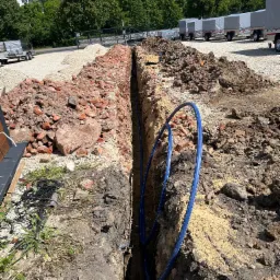 Wykopany wąski r&oacute;w w ziemi z ułożoną niebieską rurą. Po obu stronach wykopu hałdy ziemi i gruzu. W tle zaparkowane przyczepy i zieleń.