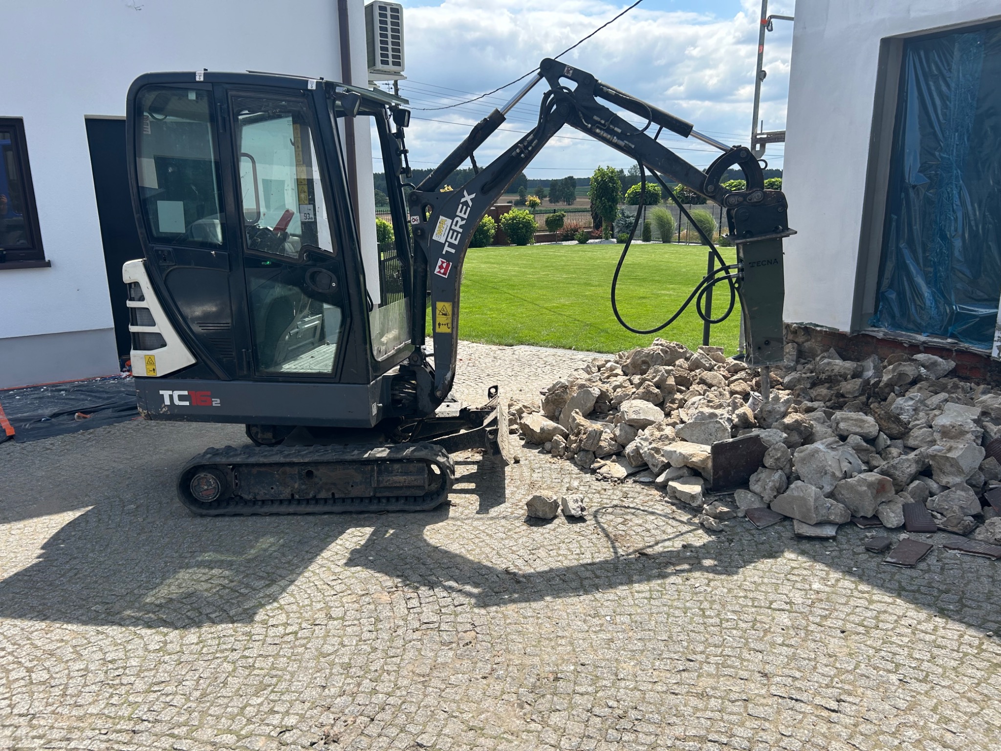 Mała koparka Terex z młotem pneumatycznym kruszy gruz przy budynku. Widoczny ogród w tle. Praca w toku, kostka brukowa na pierwszym planie.