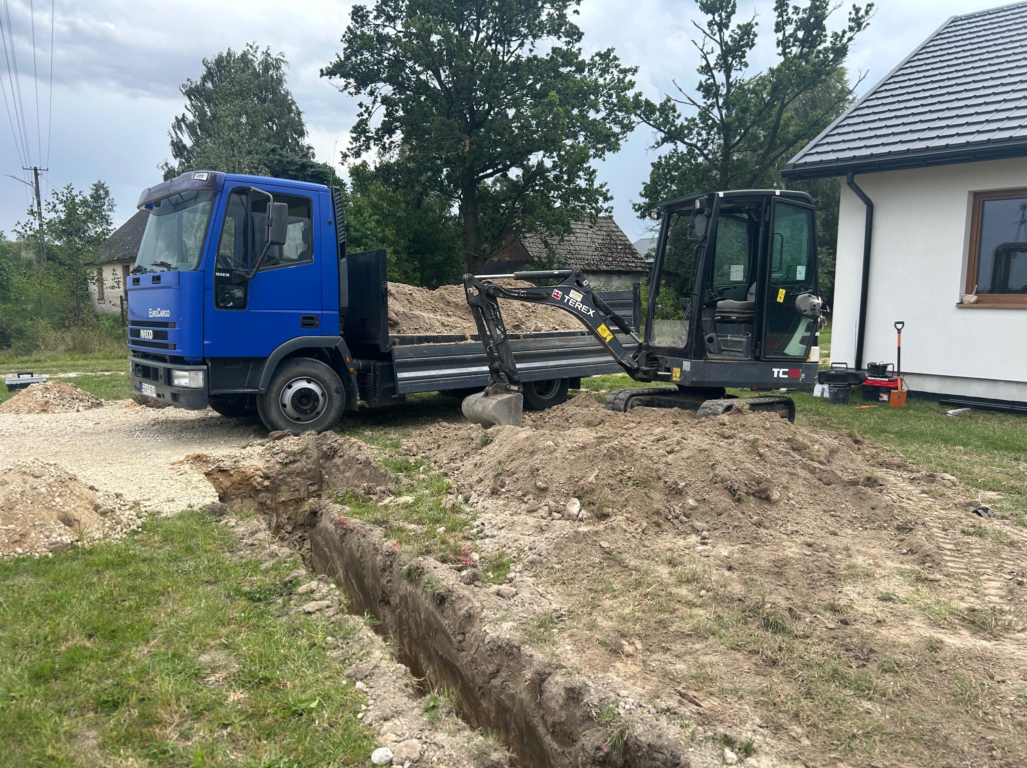 Niebieska ciężarówka z mini koparką na pace, obok wykopany wąski rów w trawie, dom w tle. Prace ziemne w toku, przygotowanie pod instalacje.