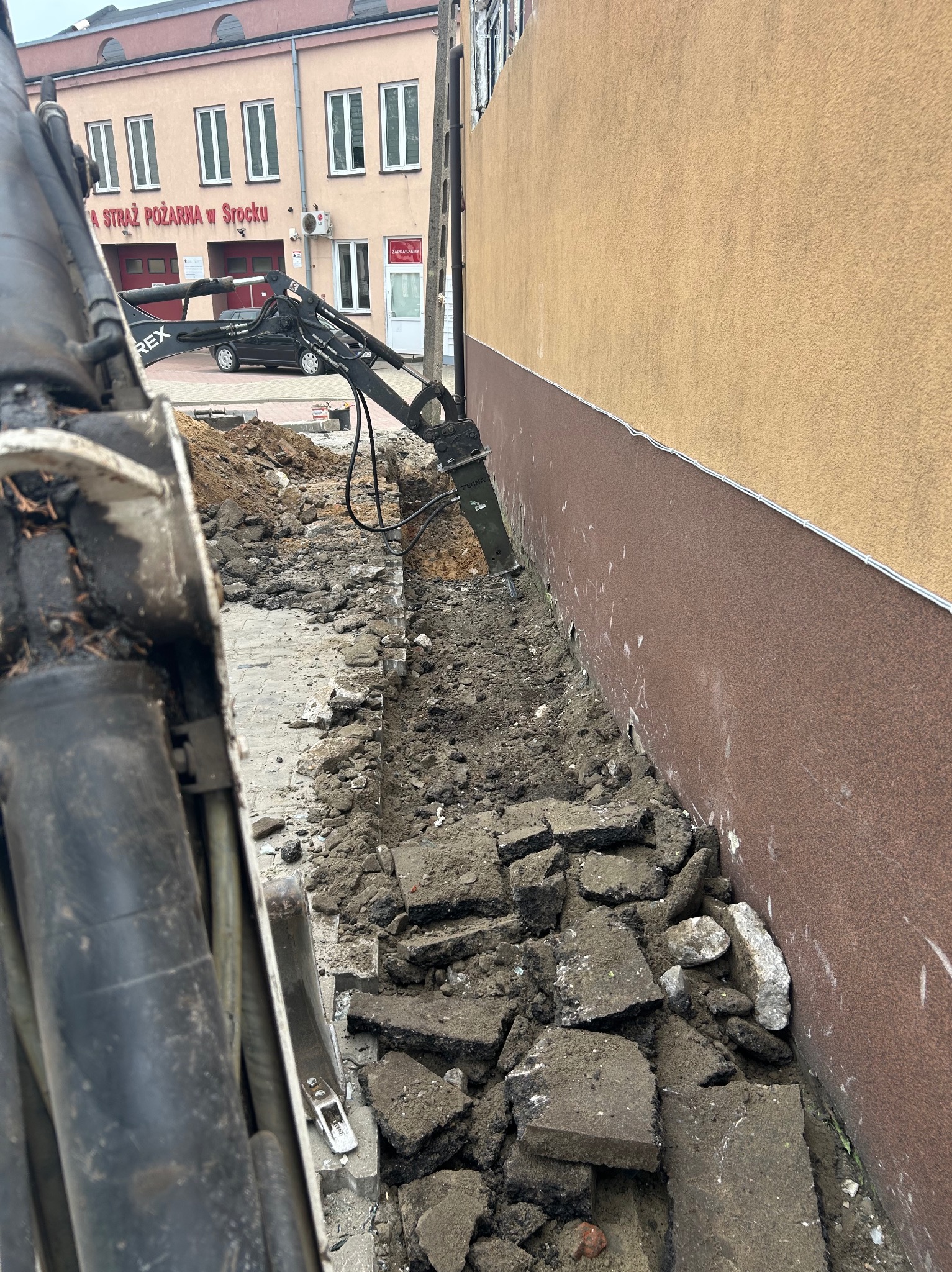Hydrauliczny młot wyburzeniowy kruszy beton przy ścianie budynku. Widoczne gruz, ziemia i maszyna budowlana. W tle budynek straży pożarnej w Srocku.