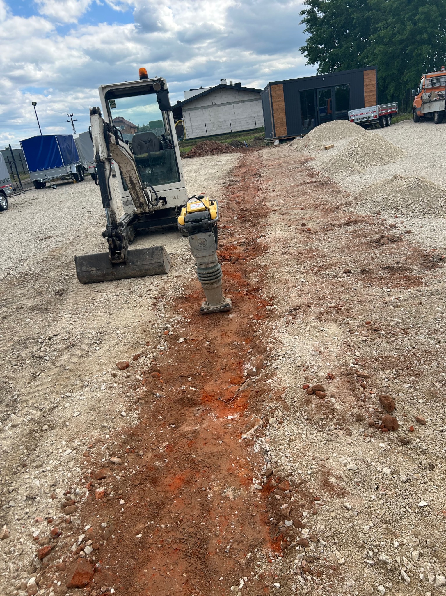 Mini koparka i ubijarka w wykopie pod instalacje na żwirowym terenie. Widoczne budynki w tle.