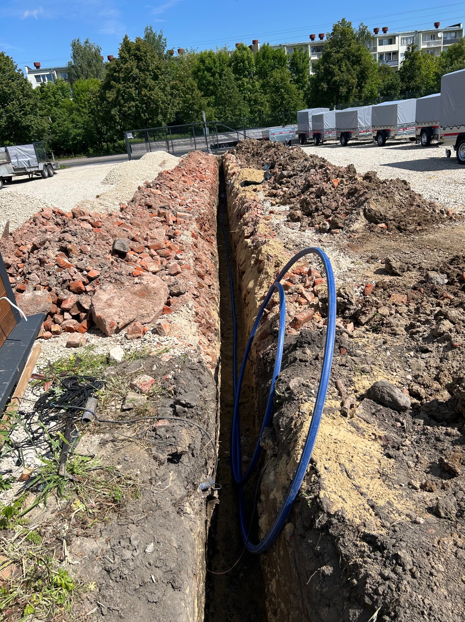 Wykopany wąski rów w ziemi z ułożoną niebieską rurą. Po obu stronach wykopu hałdy ziemi i gruzu. W tle zaparkowane przyczepy i zieleń.