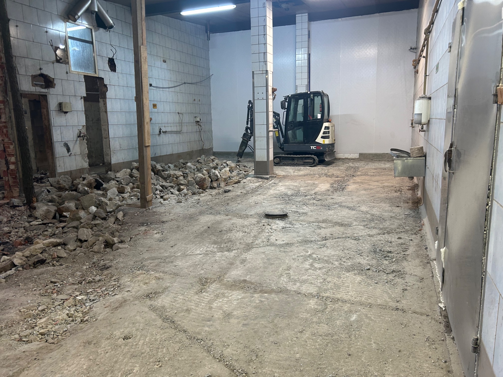 Wnętrze w trakcie rozbiórki: gruz, uszkodzone ściany z białymi płytkami, mini koparka w tle, widoczne ślady po pracach ziemnych.