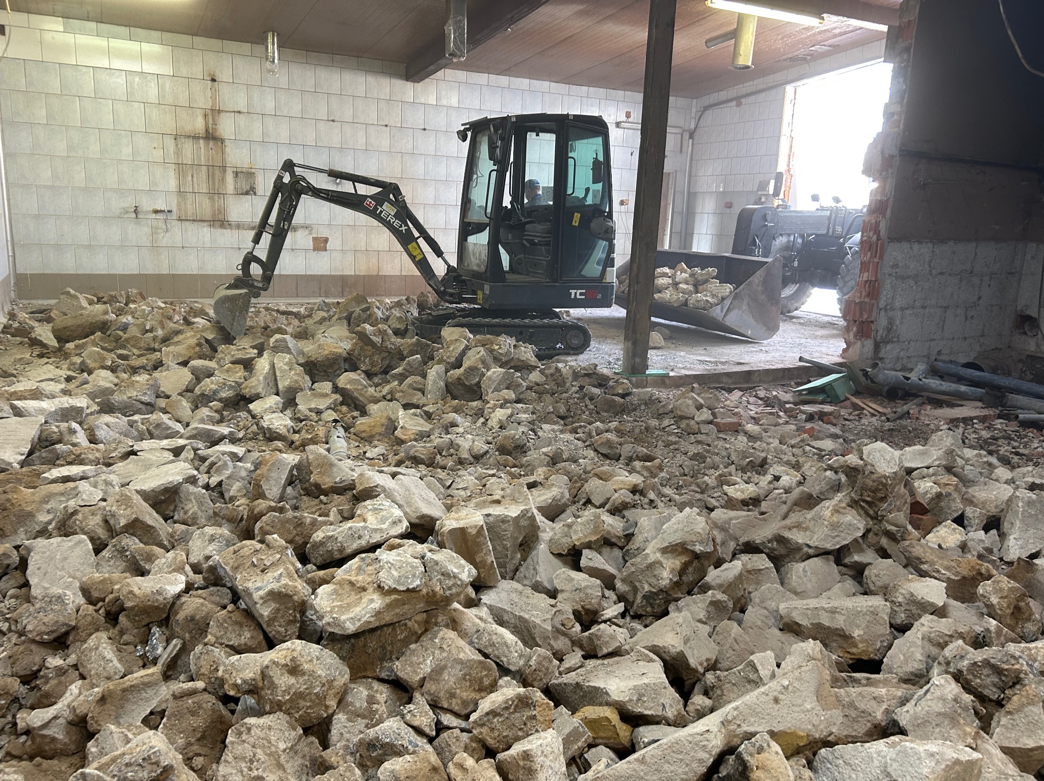 Mini koparka Terex na tle gruzu wewnątrz budynku w trakcie rozbiórki. W tle widoczny traktor z łyżką.