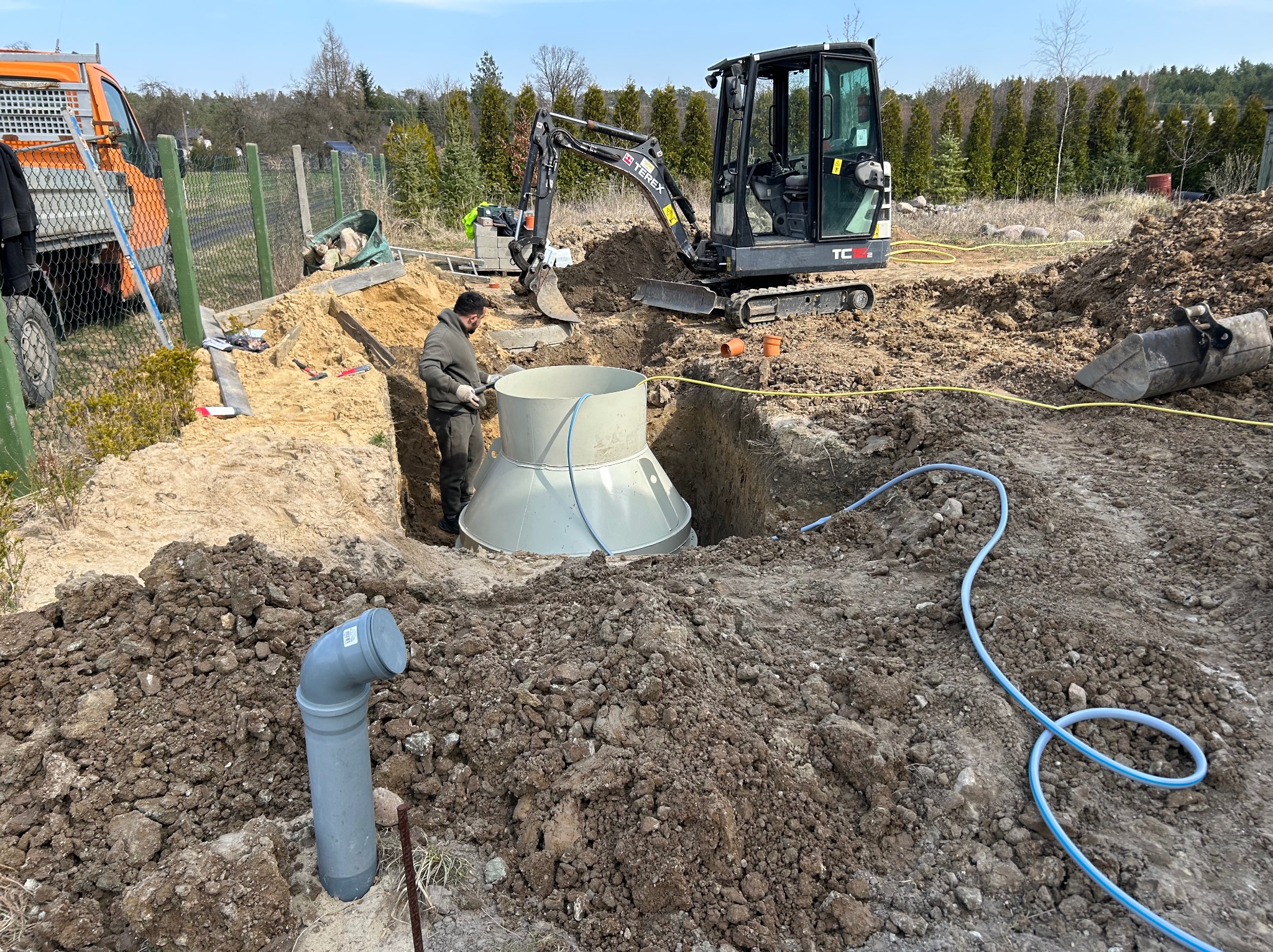 Montaż szamba: Pracownik w wykopie obok zbiornika, minikoparka w tle. Widoczny pomarańczowy samochód i zielone ogrodzenie. Niebieski przewód na ziemi.