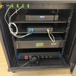 Szafa rack z zainstalowanymi urządzeniami sieciowymi: patch panel, switch, zasilacz awaryjny VOLT Polska i listwa zasilająca. Widoczne kable światłowodowe i ethernet.