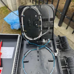 Otwarta skrzynka telekomunikacyjna z widocznymi kablami światłowodowymi i narzędziami. Układanie kabli w czarnej skrzynce na zewnątrz budynku.