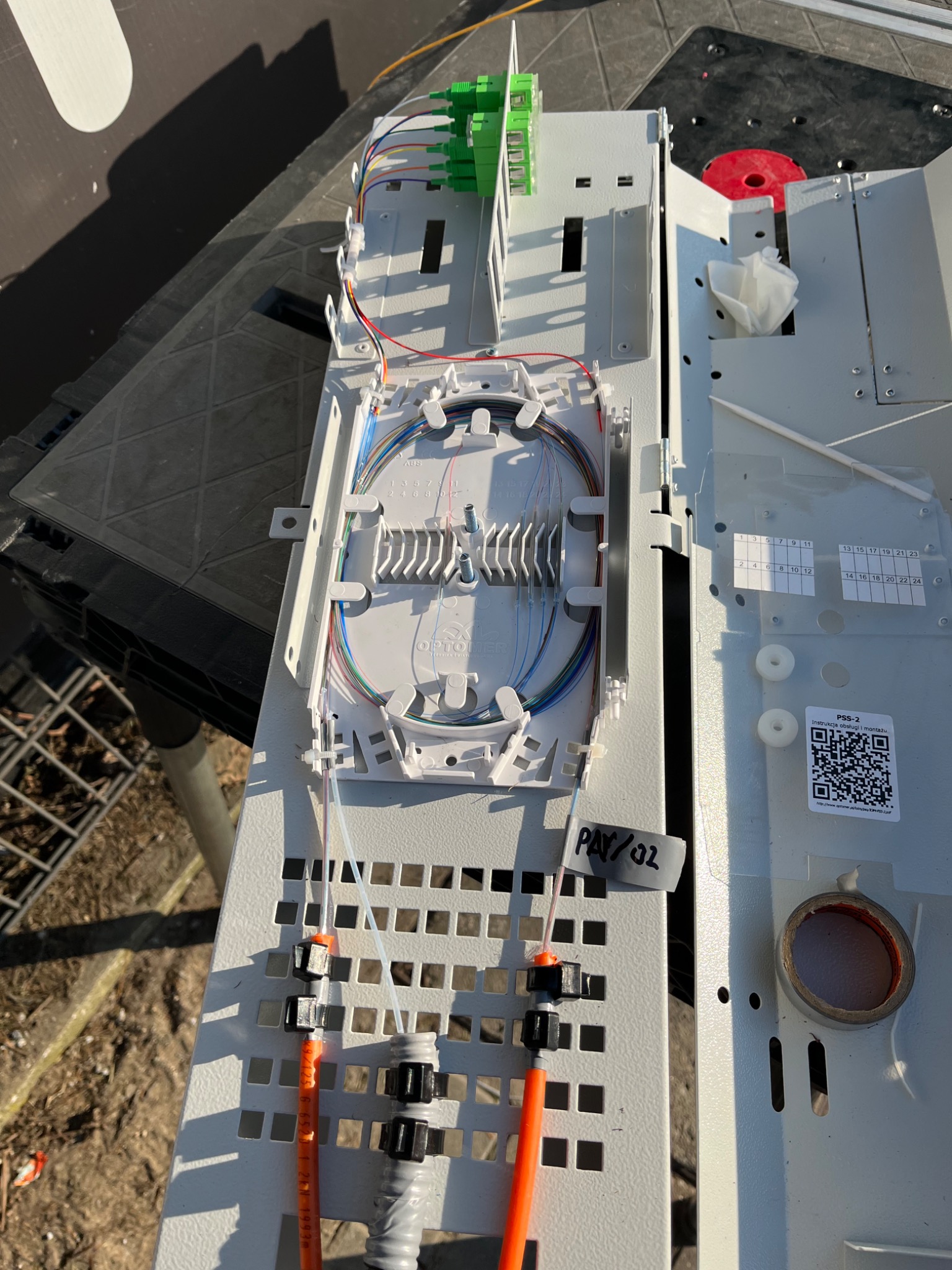 Otwarta skrzynka telekomunikacyjna z poprowadzonymi światłowodami, widoczne kable w różnych kolorach, złącza światłowodowe, etykieta 'PAY/02' i instrukcja obsługi z kodem QR na obudowie.